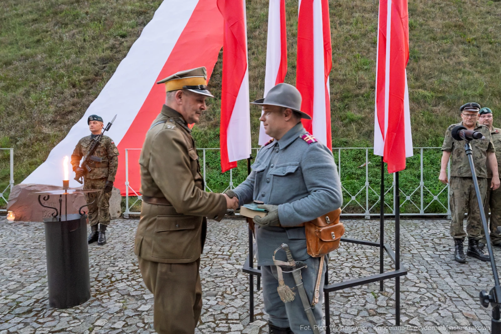 Dwaj żołnierze w mundurach, uściski dłoni, flagi narodowe w tle.  Autor: Piotr Wojnarowski
