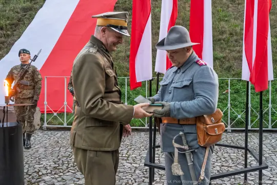 Dwaj mężczyźni w mundurach wojskowych wymieniają dokumenty, w tle flagi narodowe.