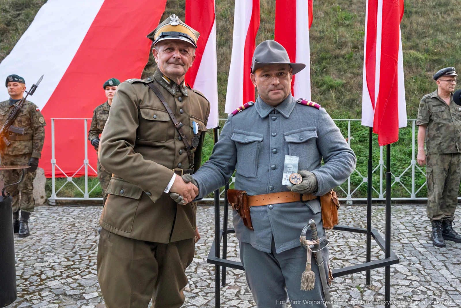 Dwaj mężczyźni w mundurach wojskowych, uściski dłoni, flagi w tle.  Autor: Piotr Wojnarowski