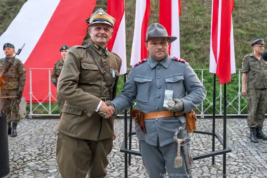 Dwaj mężczyźni w mundurach wojskowych, uściski dłoni, flagi w tle.