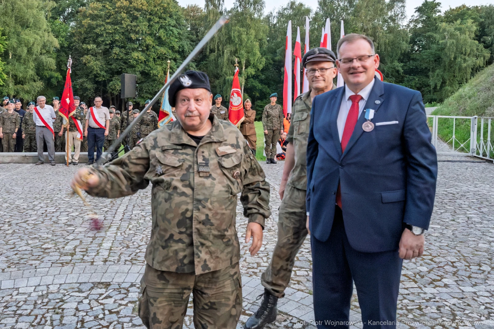 Mężczyźni w mundurach wojskowych i garniturze podczas uroczystości z flagami.  Autor: Piotr Wojnarowski
