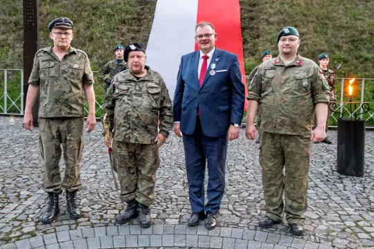 Trzech mężczyzn w mundurach wojskowych oraz jeden w garniturze, stojących przed flagą Polski.