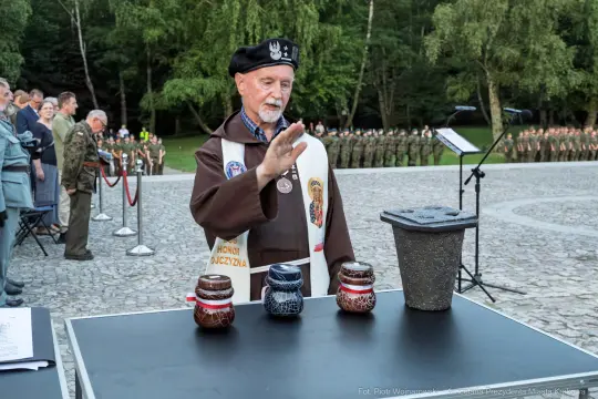 Mężczyzna w mundurze z ręką uniesioną nad trzema pojemnikami na stole. W tle zgromadzenie ludzi.