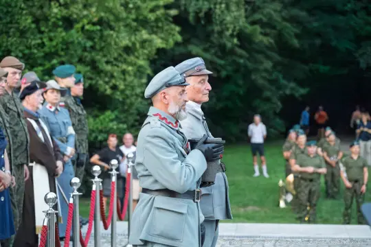 Dwaj mężczyźni w mundurach wojskowych podczas uroczystości, w tle tłum ludzi i zieleń.