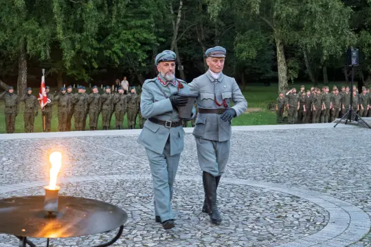 Dwaj mężczyźni w mundurach historycznych, obok żołnierze w tle, ceremonia z ogniem.