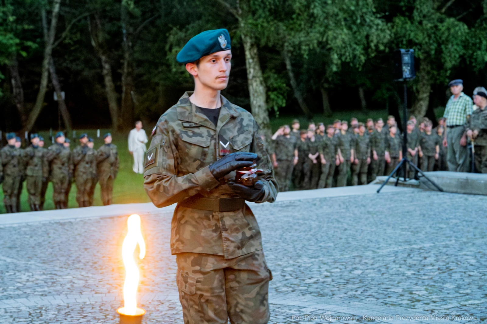 Żołnierz w mundurze z beretem, trzymający przedmiot, obok płonący znicz i grupa żołnierzy w tle.  Autor: Piotr Wojnarowski