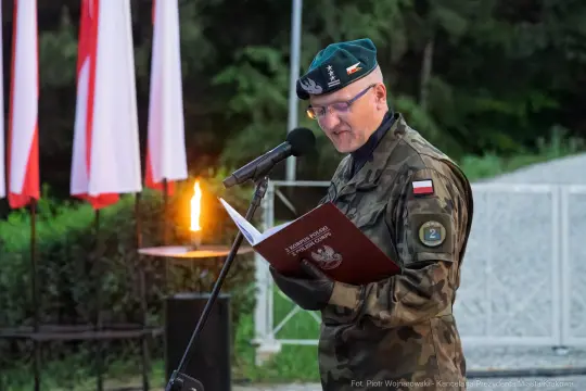 Mężczyzna w mundurze wojskowym czyta z książki podczas ceremonii, w tle flagi Polski.