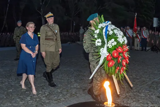 Ceremonia złożenia wieńca przez żołnierza i kobietę w mundurze, z uczestnikami w tle.