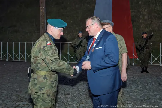Generał w mundurze wojskowym wita się z urzędnikiem w otoczeniu żołnierzy.