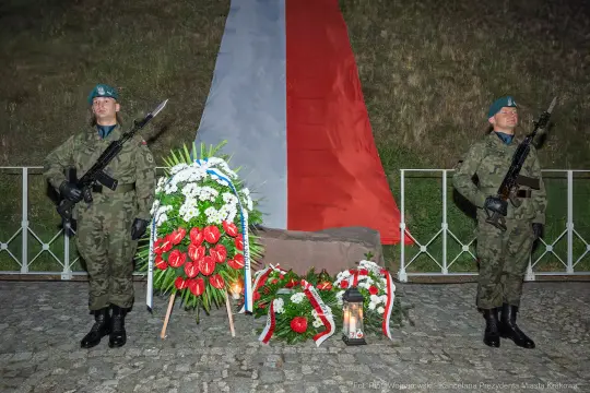 Żołnierze w mundurach stoją przy pomniku z kwiatami i zniczem, w tle flaga Polski.