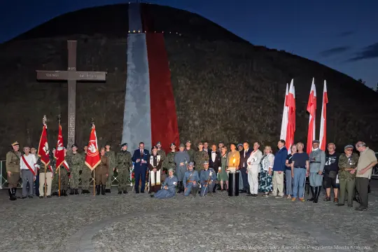 Uczestnicy uroczystości patriotycznej przy pomniku, z flagami i zniczami.