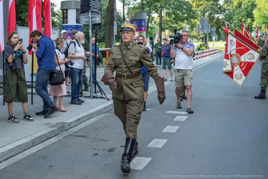 Mężczyzna w mundurze wojskowym idzie ulicą, obok flagi i grupy ludzi z kamerami.