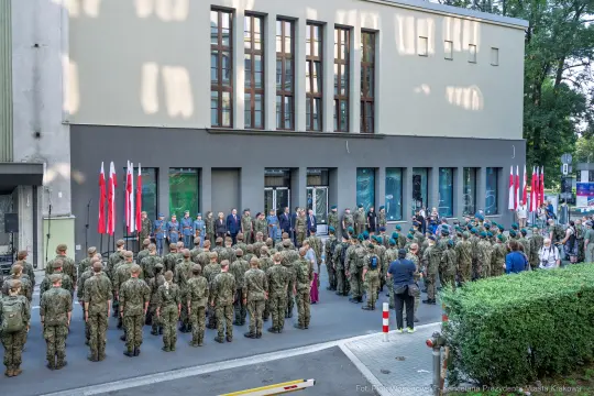 Uroczystość z udziałem żołnierzy i flag narodowych przed budynkiem.