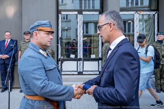 Uroczystość wręczenia odznaczeń, mężczyźni w mundurach i garniturach, uściski dłoni.