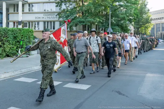 Marsz weteranów z flagą, uczestnicy w mundurach i koszulkach, ulica w Krakowie.