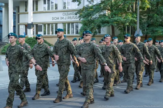 Grupa żołnierzy w mundurach maszerujących wzdłuż budynku.