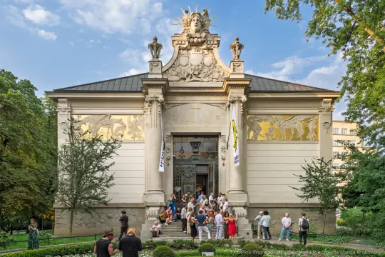 Budynek z ozdobną fasadą, ludzie na schodach, zieleń wokół.