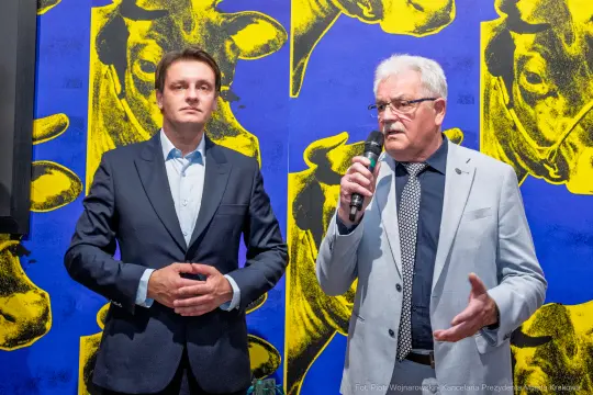 Dwaj mężczyźni podczas wystąpienia, w tle kolorowa grafika z wizerunkiem krów.