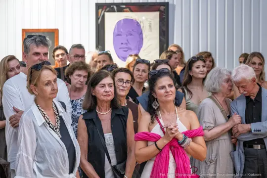 Grupa ludzi na wydarzeniu artystycznym, z portretem w tle.