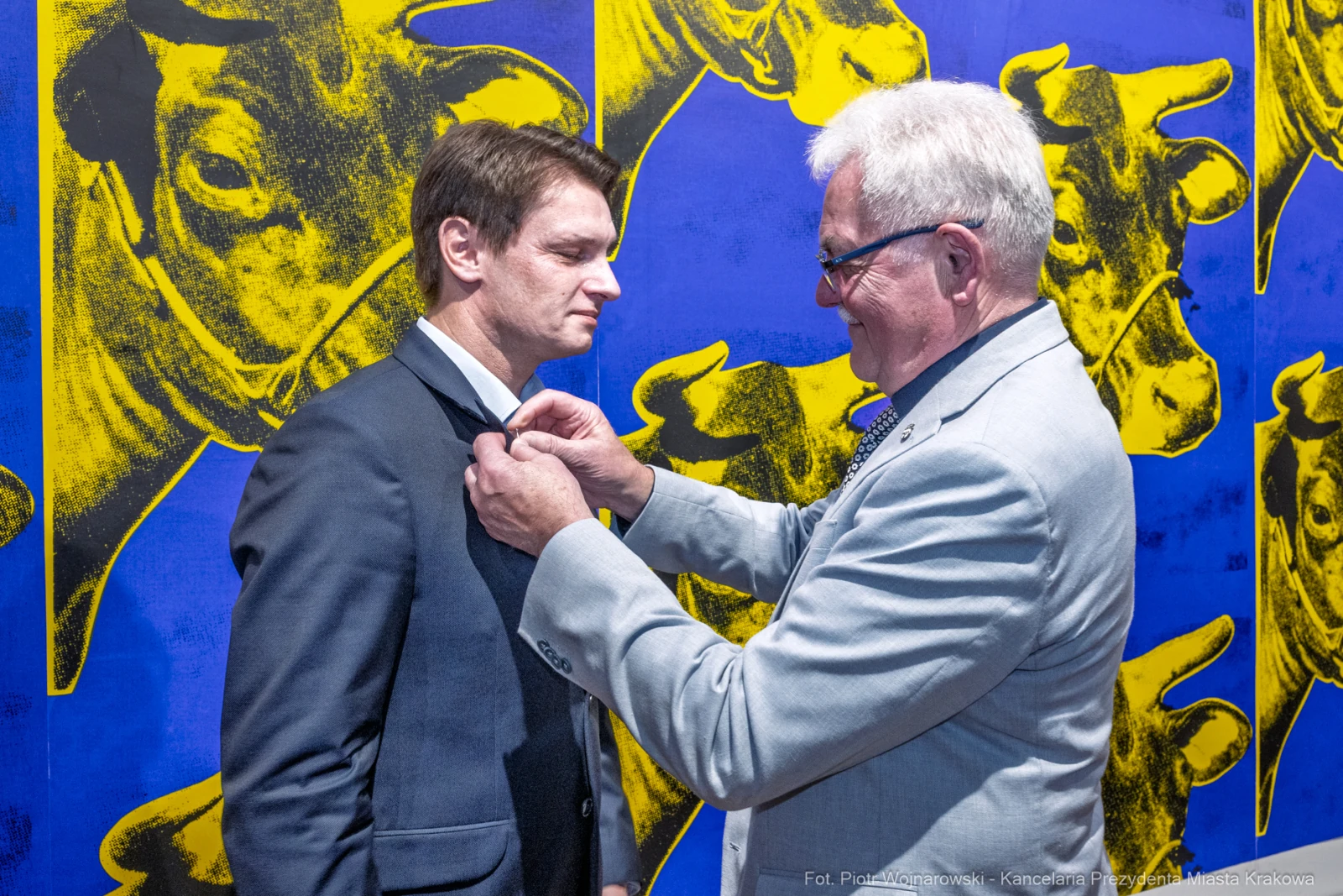 Mężczyzna w garniturze otrzymuje odznaczenie od innego mężczyzny w eleganckim stroju. Tło z kolorowymi motywami.  Autor: Piotr Wojnarowski