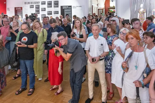 Tłum ludzi na wydarzeniu artystycznym, niektórzy trzymają aparaty i telefony.