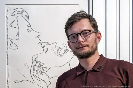 Mężczyzna w okularach stoi obok minimalistycznej grafiki przedstawiającej parę w intymnej chwili.
