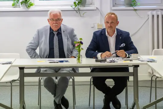 Dwaj mężczyźni siedzą przy stole, obok kwiatek w wazonie.