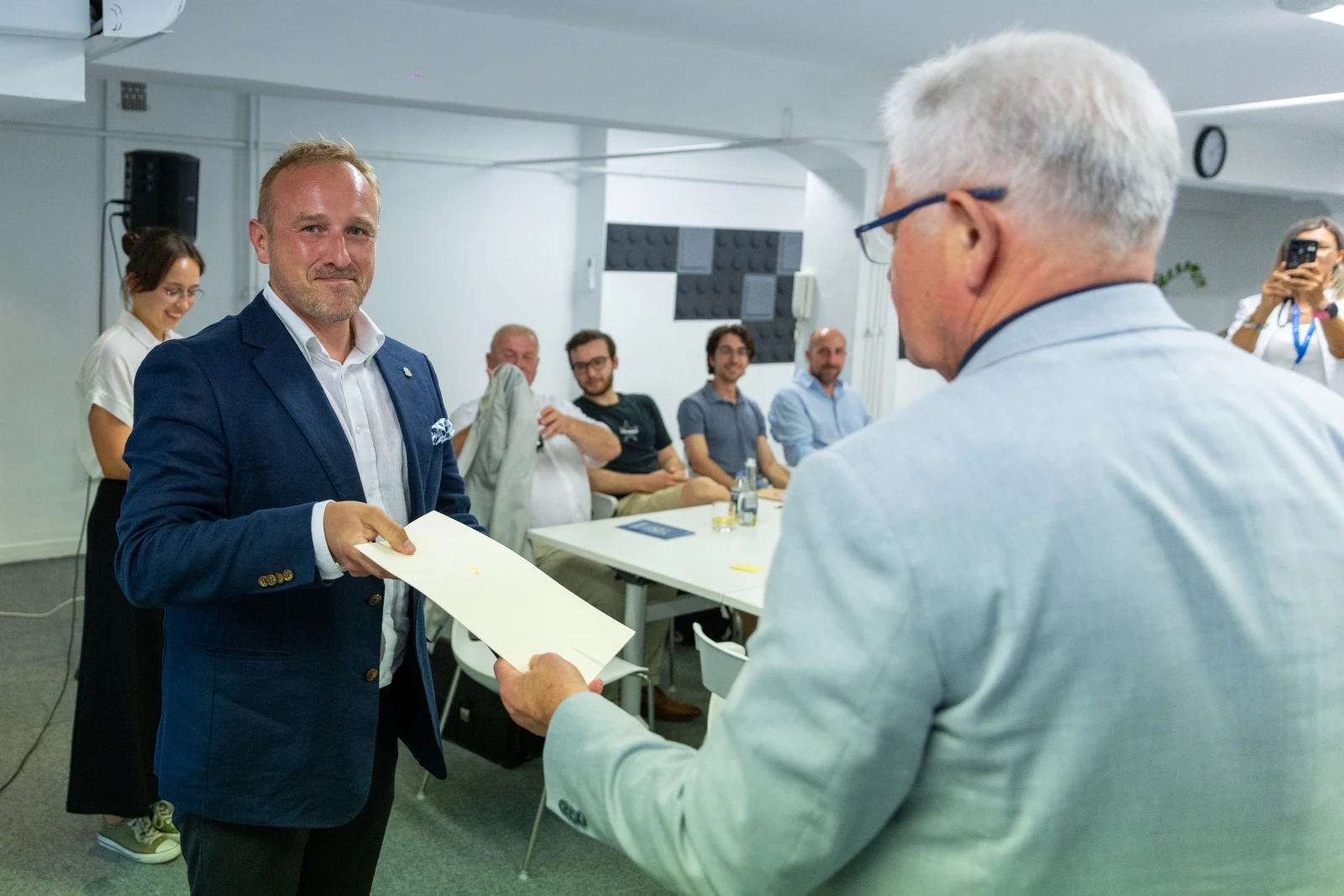 Mężczyzna w garniturze odbiera kopertę od starszego mężczyzny w jasnym garniturze. W tle grupa ludzi.  Autor: Bogusław Świerzowski