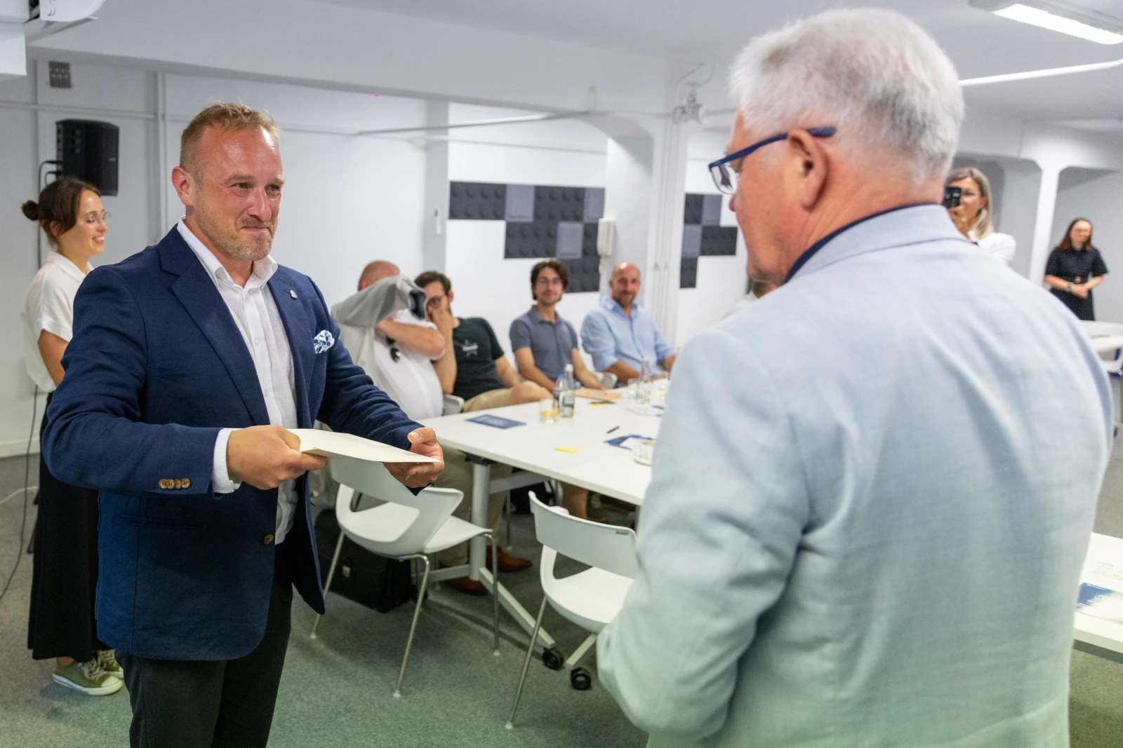 Mężczyzna w garniturze wręcza dokument innemu mężczyźnie w jasnym ubraniu. W tle grupa osób.  Autor: Bogusław Świerzowski