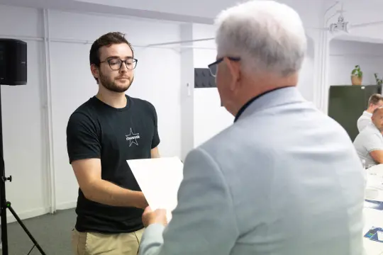 Mężczyzna w okularach wręcza dokument innemu mężczyźnie w jasnym garniturze.