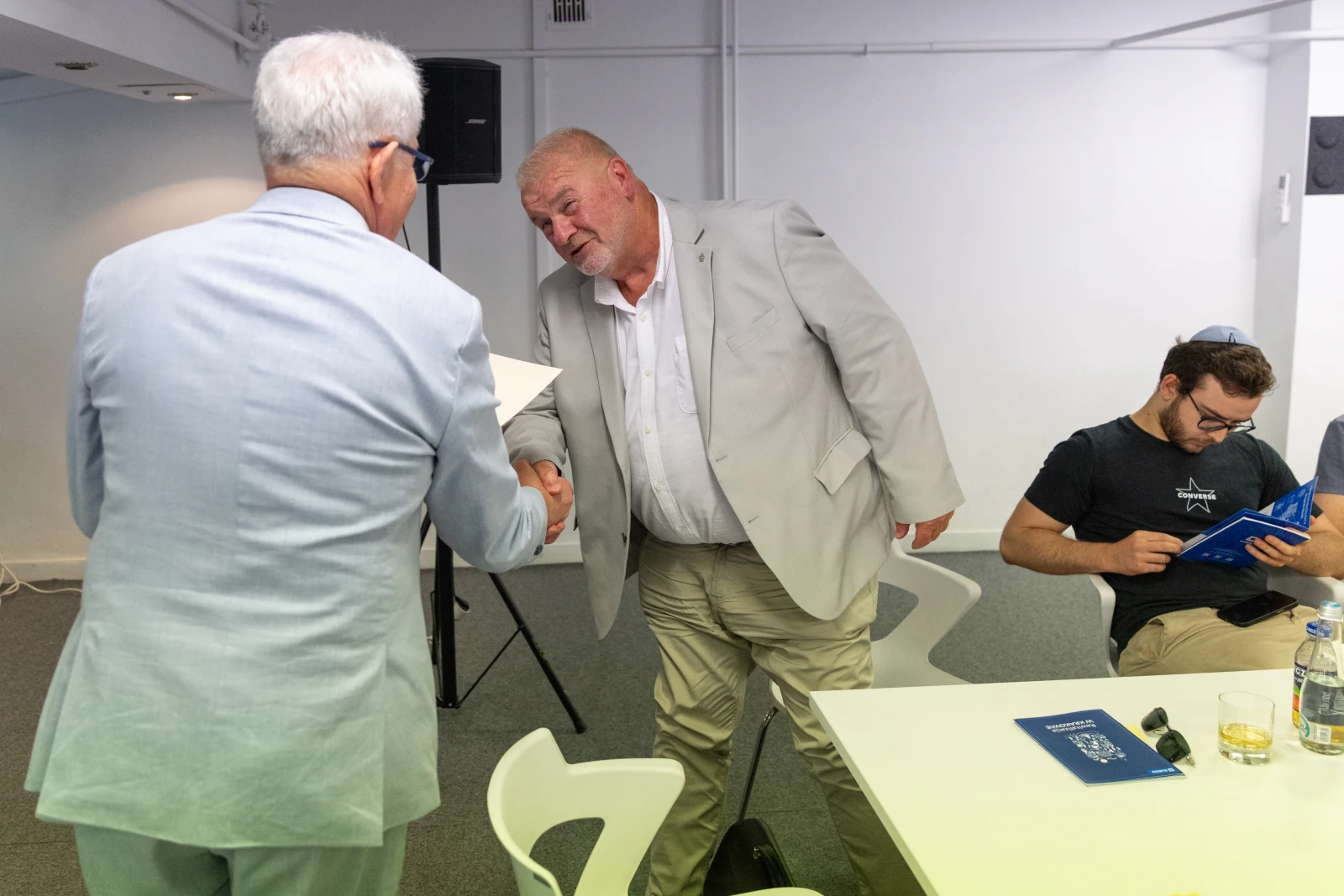 Dwaj mężczyźni podają sobie ręce, jeden z nich w jasnym garniturze, drugi w szarym. W tle siedzi mężczyzna z książką.  Autor: Bogusław Świerzowski