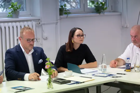 Spotkanie w biurze, uczestnicy przy stole, kobieta w okularach, kwiat w wazonie.