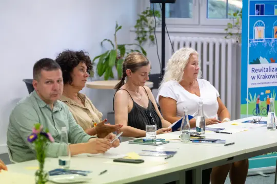 Cztery osoby siedzące przy stole, uczestniczące w spotkaniu, z notatkami i szklankami wody.