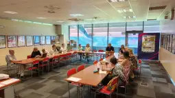 Spotkanie grupy ludzi w sali konferencyjnej z dużymi oknami i stołami ustawionymi w układzie U.
