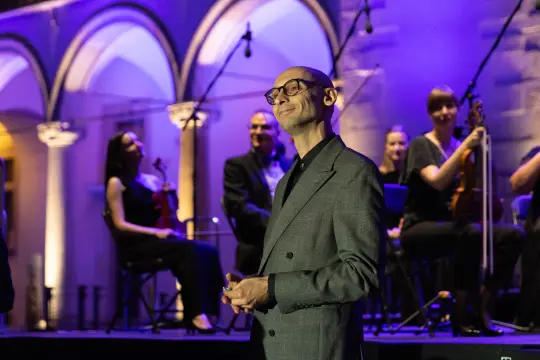 Mężczyzna w okularach na tle muzyków grających na instrumentach w oświetlonym wnętrzu.