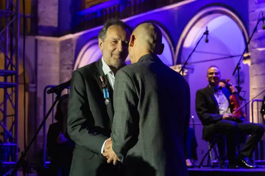 Uścisk dłoni dwóch mężczyzn podczas ceremonii, w tle muzycy na scenie.