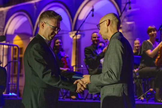 Dwóch mężczyzn uścisnęło sobie dłonie podczas ceremonii wręczenia nagrody. W tle muzycy.