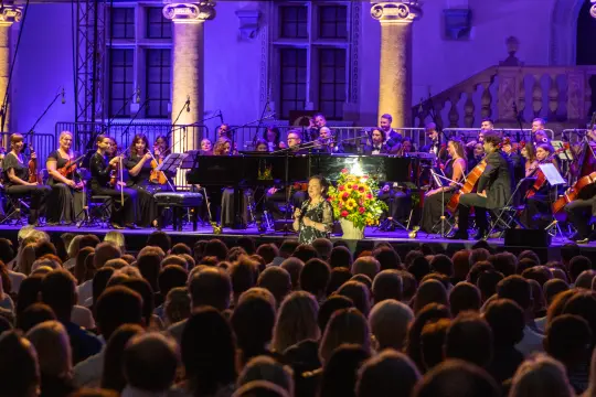 Koncert z orkiestrą, artystka śpiewająca przed publicznością, kwiaty na scenie.