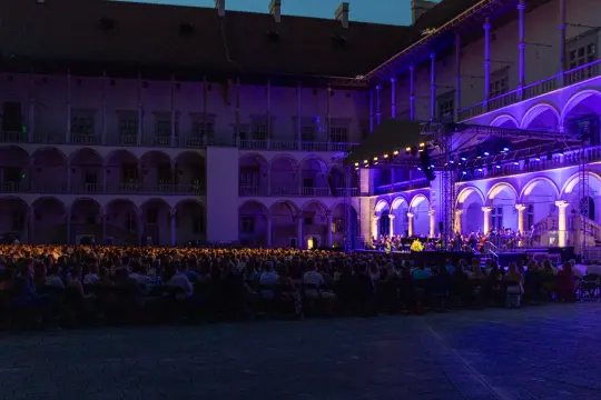 Koncert na świeżym powietrzu w otoczeniu historycznej architektury, oświetlony niebieskimi światłami.