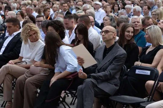 Grupa ludzi siedzących na widowni, niektórzy trzymają kartki, w tle widać tłum.