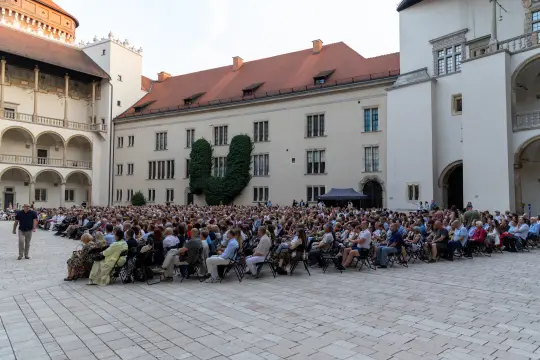 Tłum ludzi siedzących na dziedzińcu, uczestniczących w wydarzeniu na świeżym powietrzu.