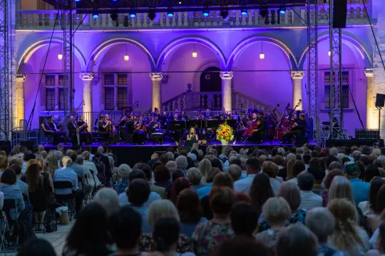 Koncert orkiestry na tle oświetlonych budynków, z widownią siedzącą przed sceną.