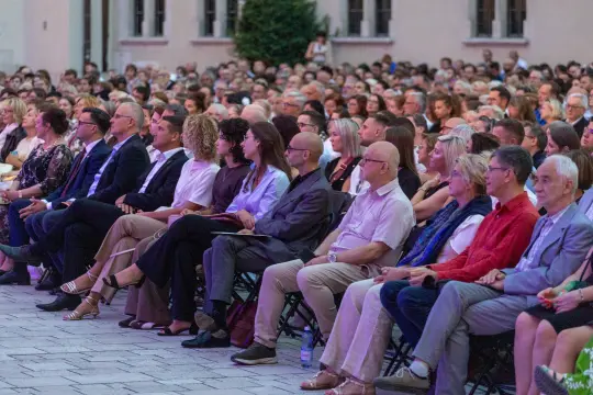 Tłum ludzi siedzących na krzesłach, uczestniczących w wydarzeniu na świeżym powietrzu.