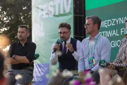 Trzech mężczyzn siedzi na scenie, jeden z nich mówi do mikrofonu podczas festiwalu filmowego.