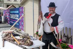 Mężczyzna w tradycyjnym stroju ludowym trzyma instrumenty muzyczne i stoi przy stole z rzemiosłem.