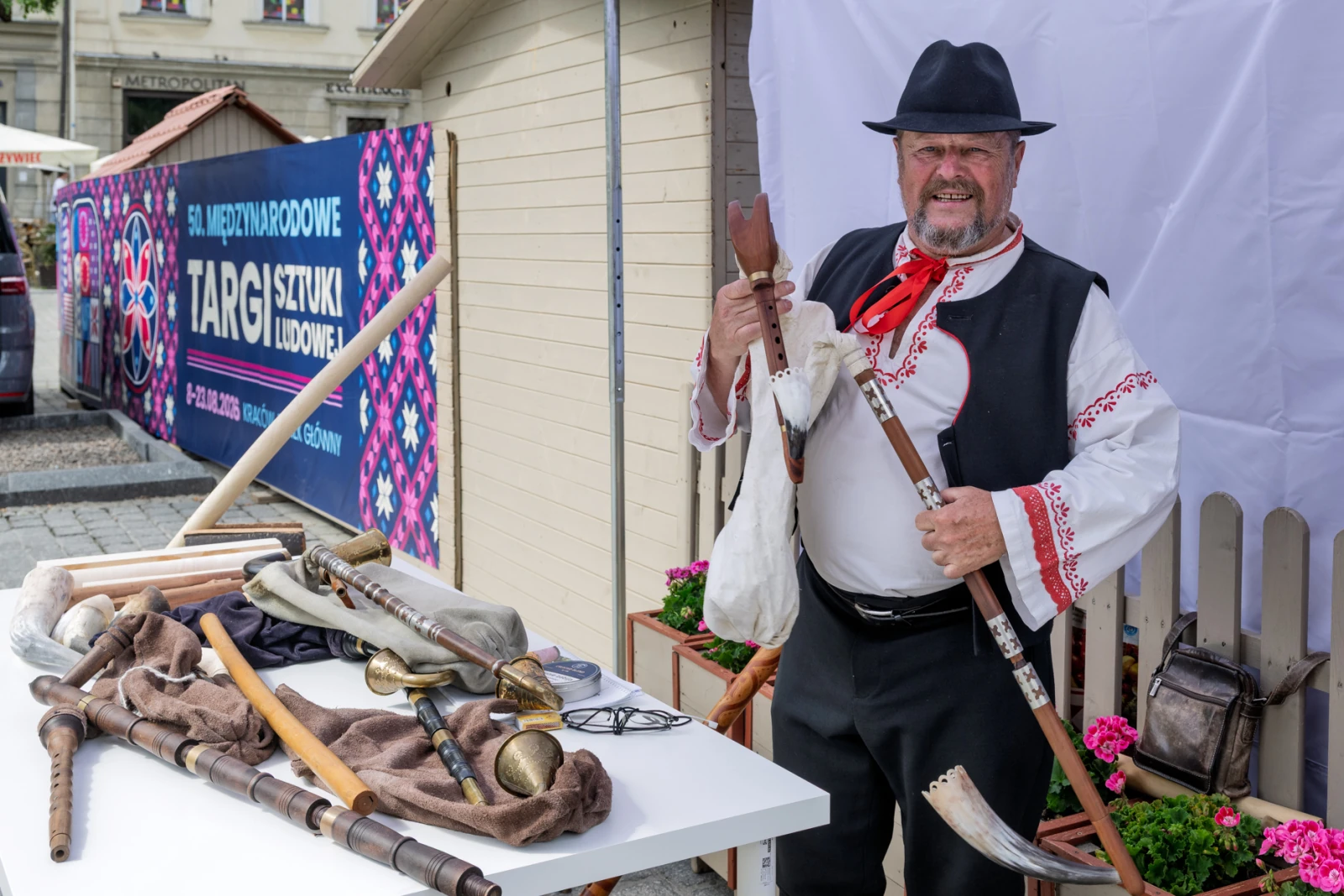 Mężczyzna w tradycyjnym stroju ludowym trzyma instrumenty muzyczne i stoi przy stole z rzemiosłem.  Autor: Piotr Wojnarowski