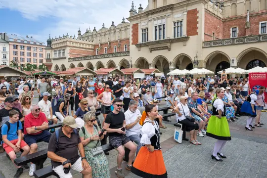 Tłum ludzi na rynku, występy artystyczne, kolorowe stroje, letnia atmosfera.