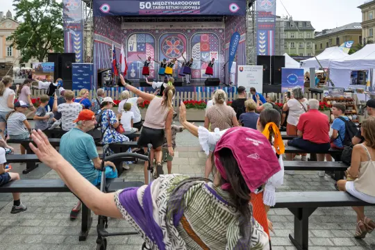 Tłum ludzi na festiwalu, występy artystyczne na scenie, kolorowe stroje i radosna atmosfera.