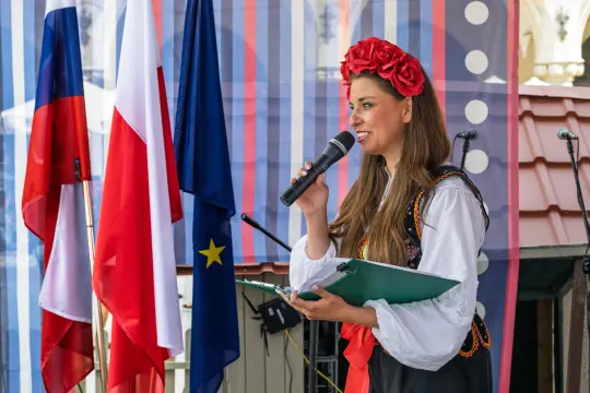 Kobieta w tradycyjnym stroju ludowym trzyma mikrofon i notatnik, przemawia na tle flag.
