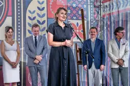 Kobieta w czarnej sukience przemawia do zgromadzenia, obok niej mężczyźni w garniturach.